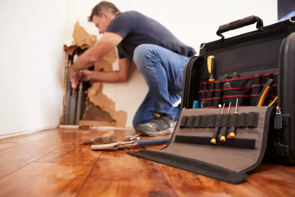 Relief Remediation technician repairing a wall behind a burst pipe with a tool kit open on the floor